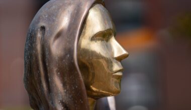 Seitenansicht einer Metallstatue, die Satoshi Nakomoto mit einem menschlichen Gesicht mit Kapuze darstellt, mit einer glatten, reflektierenden goldenen Oberfläche und einer bronzefarbenen Kapuze.