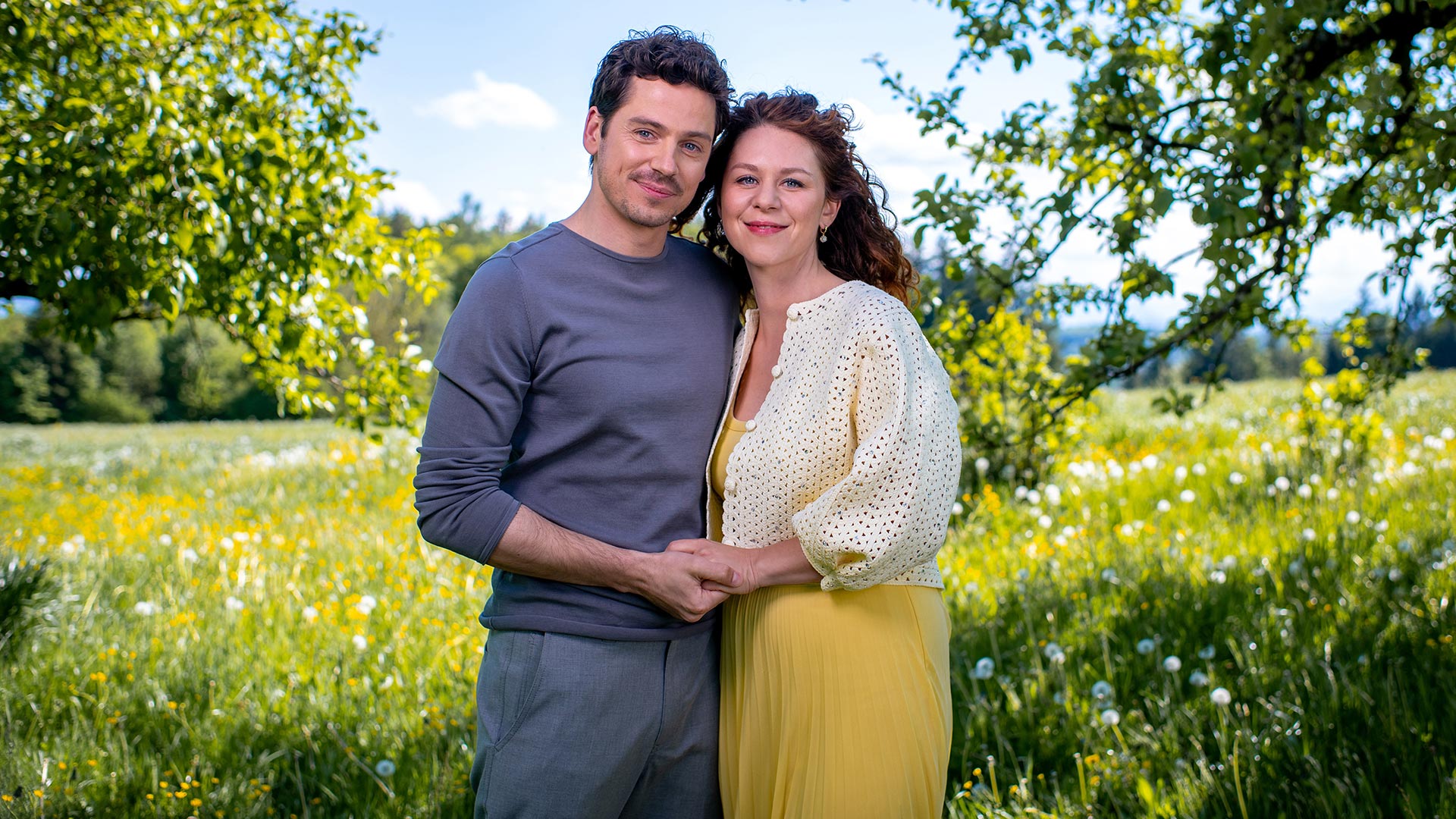 Auf dem Bild zu Sturm der Liebe Staffel 22 ist das neue Traumpaar der Staffel in einer sommerlichen Wiesenlandschaft zu sehen. Die beiden stehen eng beieinander, halten sich an den Händen und lächeln in die Kamera.