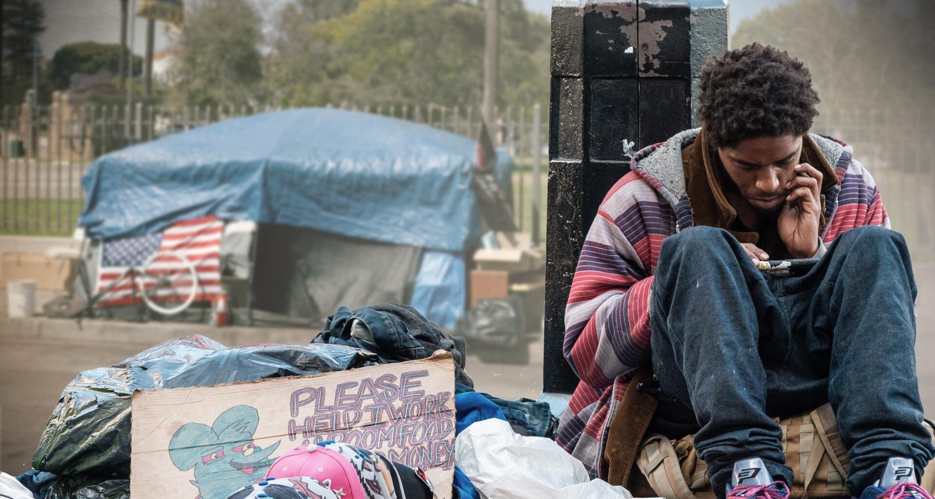 USA extrem: Obdachlos in Los Angeles": in der linken Bildhälfte improvisiertes Zelt aus Planen mit US-Fahne vor einem Zaun vor parkähnlichem Grundstück im Hintergrund, davor im Vordergrund Müllsäcke und Pappschild mit Schriftzug PLEASE HELP I WORK FOR ROOM FOOD MONEY und in der rechten Bildhälfte ein junger Mann, der auf einer Tasche sitzt, am unteren Teil einer Straßenlaterne lehnt und auf sein Smartphone schaut.