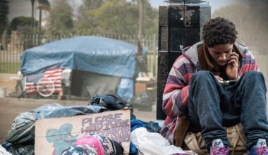 USA extrem: Obdachlos in Los Angeles": in der linken Bildhälfte improvisiertes Zelt aus Planen mit US-Fahne vor einem Zaun vor parkähnlichem Grundstück im Hintergrund, davor im Vordergrund Müllsäcke und Pappschild mit Schriftzug PLEASE HELP I WORK FOR ROOM FOOD MONEY und in der rechten Bildhälfte ein junger Mann, der auf einer Tasche sitzt, am unteren Teil einer Straßenlaterne lehnt und auf sein Smartphone schaut.