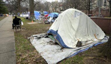 Mehrere Zelte stehen auf einem Stück Wiese. Dahinter sind Häuser zu sehen. Vor den Zelten steht eine Park-Bank. Darauf sitzt ein Mann.