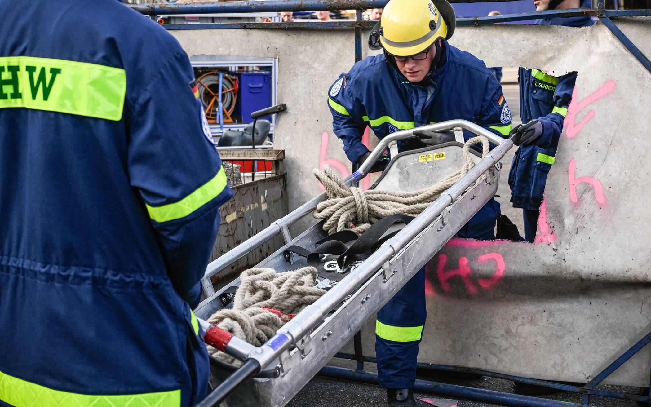  Helfer des THW in Schutzkleidung reichen eine Schleifkorbtrage durch eine Öffnung in einem Blech. 