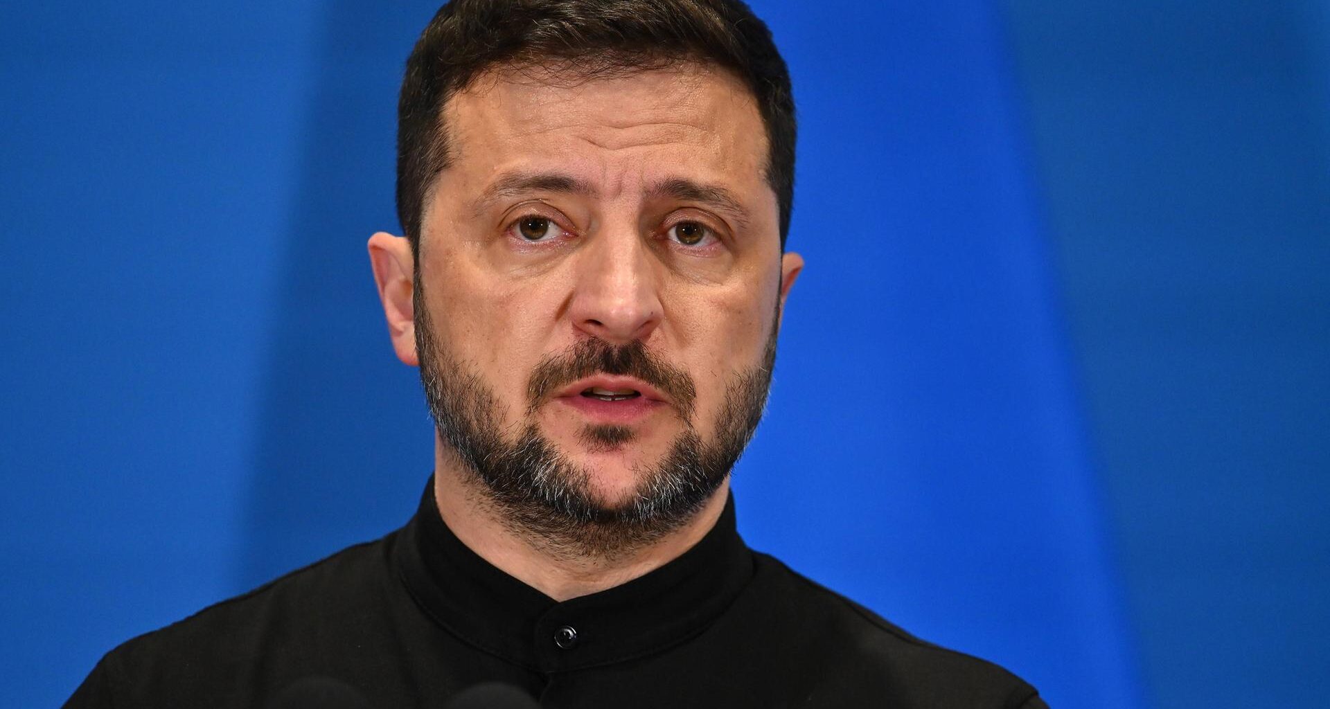 Der ukrainische Präsident Wolodymyr Selenskyj steht vor einer blauen Wand.