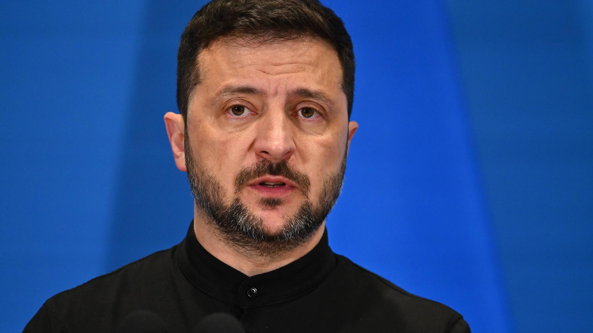 Der ukrainische Präsident Wolodymyr Selenskyj steht vor einer blauen Wand. 