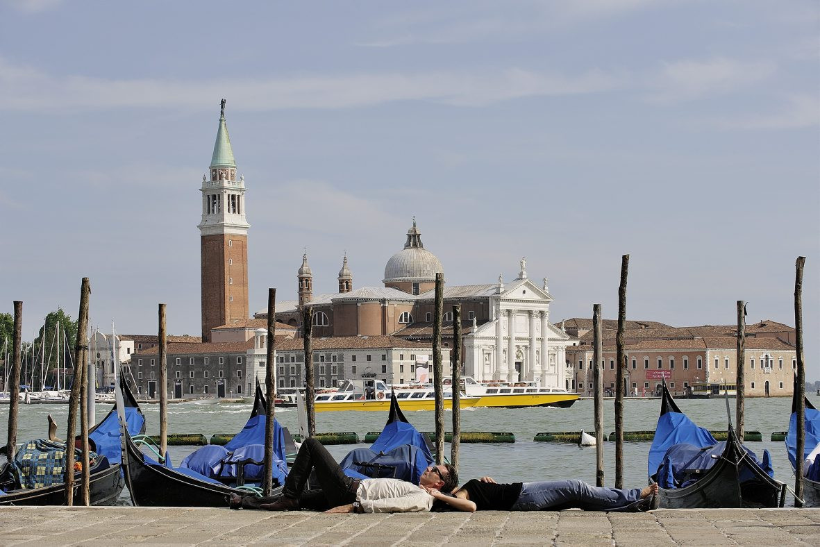 Zwei Personen entspannen auf dem Boden der Piazza San Marco in Venedig, mit San Giorgio Maggiore im Hintergrund und Gondeln im Vordergrund.