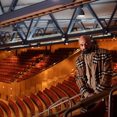 Xatar steht im leeren Zuschauerraum der Kölner Philharmonie. Er trägt ein helles kariertes Jackett.