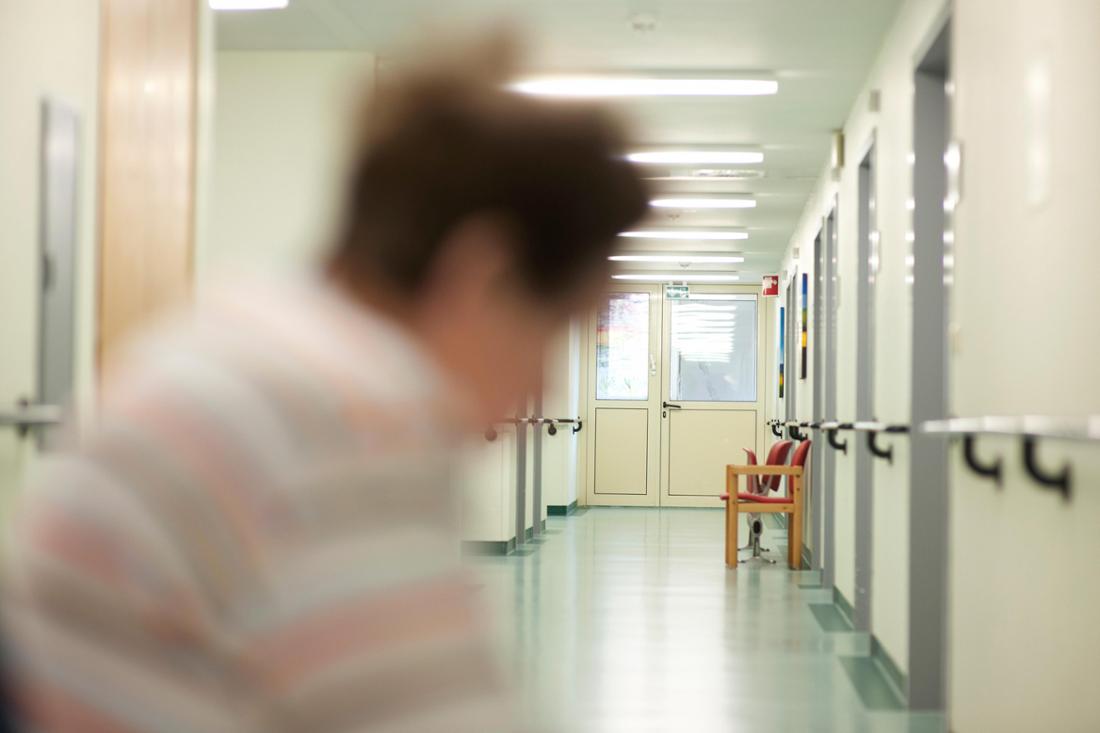 Das Foto steht symbolisch für eine psychiatrische Station. Eine Frau, ihre Erscheinung etwas verschwommen, sitzt in einem leeren Klinikflur. Im  Hintergrund stehen zwei leere rote Stühle. 