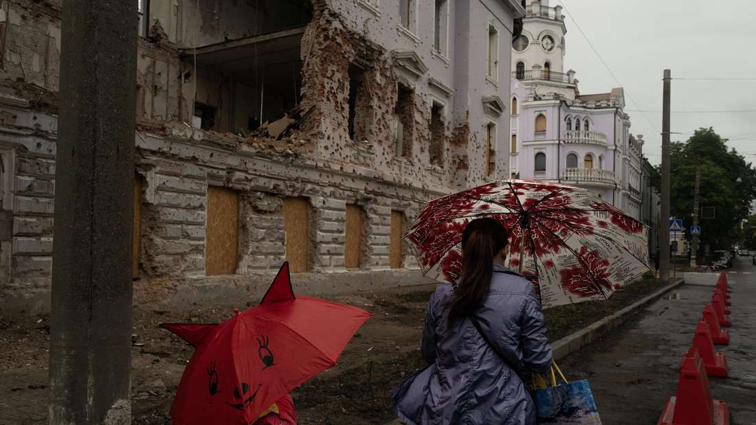 Frau und Kind mit Regenschirmen vor einem teilzerstörten alten Gebäude.