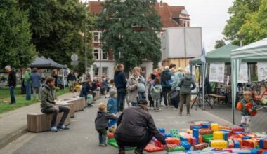Auf einer Straße stehen Menschen und unterhalten sich, dazwischen Kinder. Rechts stehen offene Zelte. Vorn liegen überdimensionale Legosteine auf dem Boden, ein Mann hockt davor, zwei Kinder halten Steine in den Händen.
