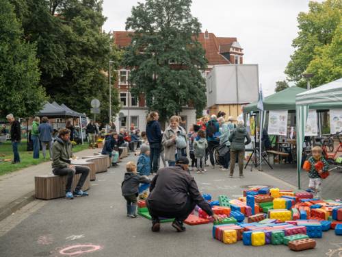 Auf einer Straße stehen Menschen und unterhalten sich, dazwischen Kinder. Rechts stehen offene Zelte. Vorn liegen überdimensionale Legosteine auf dem Boden, ein Mann hockt davor, zwei Kinder halten Steine in den Händen.