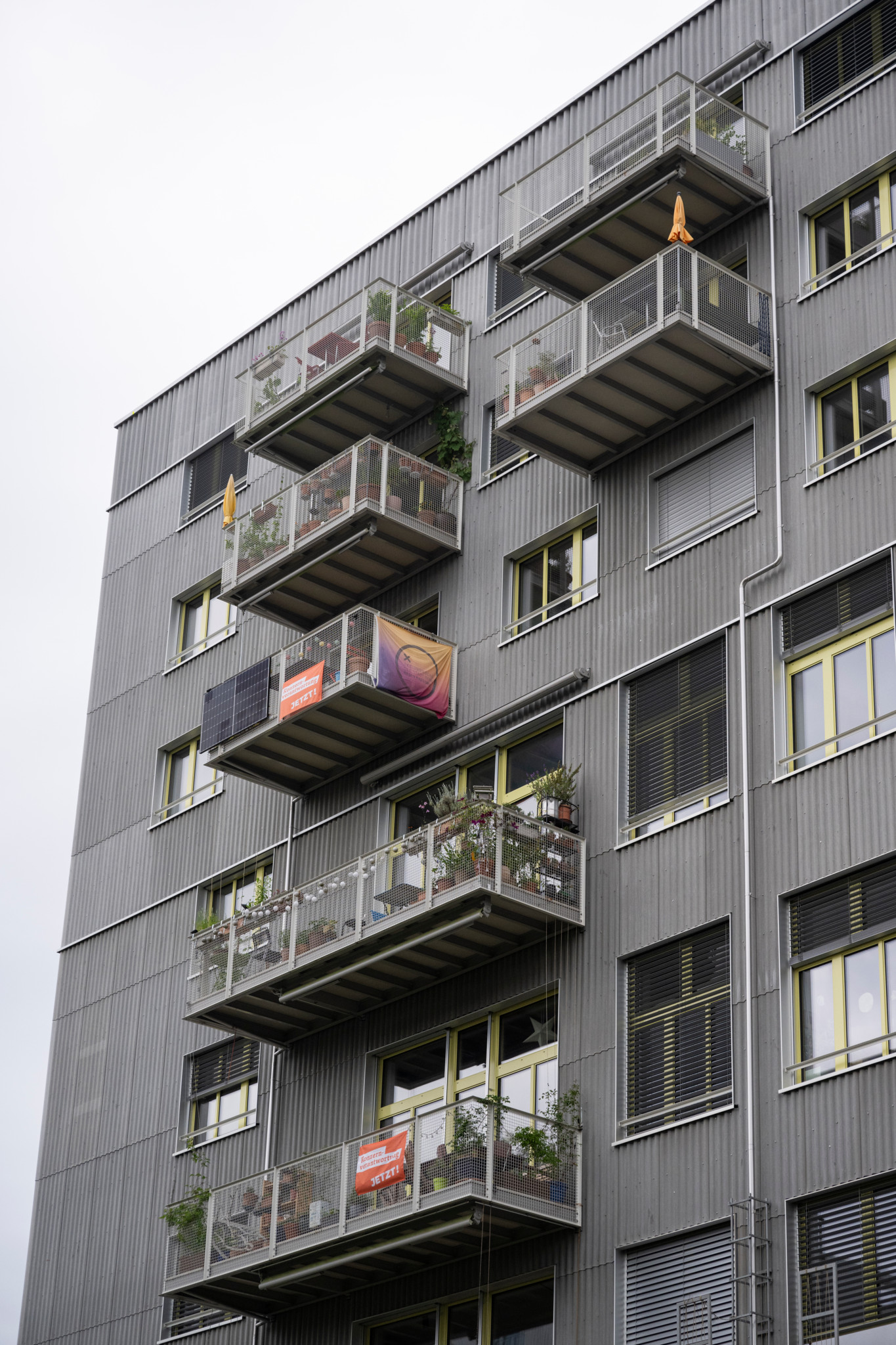 Ein modernes Wohngebäude in Bern mit mehreren Balkonen, auf denen Pflanzen stehen. Ein Balkon zeigt ein Banner mit einem Logo.