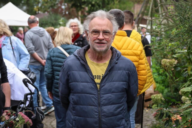 Imker Ralf Schmidt steht auf dem Gelände seiner Imkerei in Dortmund-Sölde. Im Hintergrund sind Besucher des Hoffestes zu sehen. Der Imker lächelt in die Kamera.