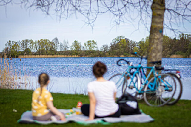 Eine Frau mit Kind sitzt am Ufer eines Sees. Fahrräder lehnen an einem Baum.