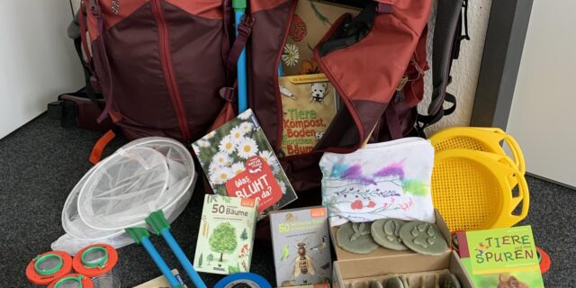 Auf der Abbildung ist ein Rucksack und verschiedene Gegenstände zum Thema Natur zu sehen, darunter Bücher, Lupen, Pinsel, eine Pinzette und ein Stethoskop. Ebenfalls zu sehen sind Netze und eine Box mit Abdrücken.