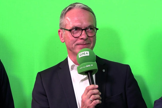 André Dora mit einem Mikrofon in der Hand vor einer grünen Wand.