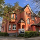 Übers Eck fotografierte Villa in der Farbe pompejanisch-rot, rechts ein kleiner Anbau in gelbem Farbton.