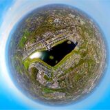 Ein sphärisches Luftbild zeigt einen See, der von Wohn- und Gewerbegebieten umgeben ist. Der Himmel ist blau und mit einigen Wolken bedeckt.