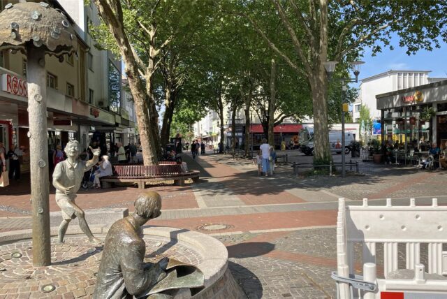 Die Fußgängerzone von Dortmund-Hombruch an einem sonnigen Tag. Im Vordergrund befindet sich ein Brunnen mit Statuen, während im Hintergrund Passanten auf dem mit Bäumen gesäumten Weg flanieren und ein Café besucht wird.