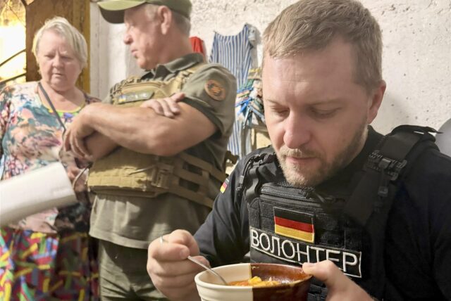 Ein Mann ist eine Suppe, er hat eine schusssichere Weste an. Daneben steht ein Mann in Uniform und eine ältere Frau