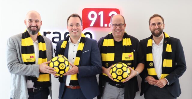 Vier Männer posieren vor dem Logo von Radio 91.2. Sie tragen Schals in den Vereinsfarben von Borussia Dortmund und halten Fußbälle mit dem BVB-Logo.