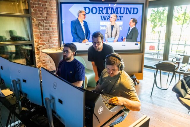 Auf einem großen Bildschirm ist zu sehen, wie zwei Journalisten den Oberbürgermeister von Dortmund, Thomas Westphal, interviewen. Davon sitzen, an einem Mischpult und vor mehreren Bildschirmen, Video-Techniker.