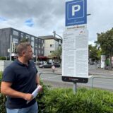 Ein Mann steht vor einem Parkschild, das mit „3x Kundenparkplätze SunsetLounge“ gekennzeichnet ist. Auf dem Schild befinden sich außerdem Hinweise zum Datenschutz und allgemeine Geschäftsbedingungen. Im Hintergrund sind Gebäude und eine Straße mit Passanten zu sehen.
