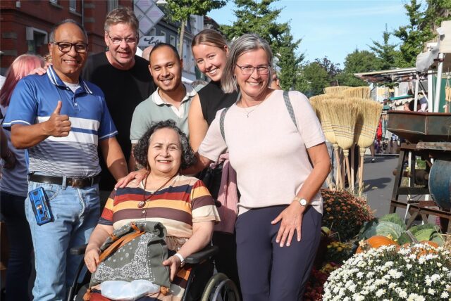 Sechs Männer und Frauen stehen beim Aplerbecker Apfelmarkt 2025 neben Blumen und Besen. Eine der Frauen sitzt im Rollstuhl. Alle lächeln in die Kamera. Einer der Männer zeigt den Daumen nach oben.