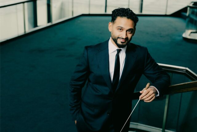 Generalmusikdirektor der Stadt Dortmund, Jordan de Souza trägt einen Anzug und blickt lächelnd in die Kamera. Im Hintergrund ist ein blauer Teppich zu sehen.