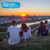 Eine Gruppe von Menschen sitzt auf einem Hügel und blickt auf eine Stadt während des Sonnenuntergangs. Im Hintergrund sind Gebäude, ein Fluss und ein Fernsehturm zu sehen.