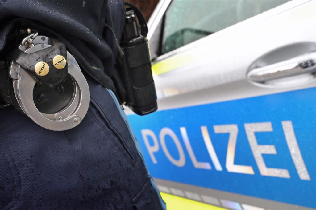 Ein Polizist mit Handschellen und einer Pistole am Gürtel steht vor einem Streifenwagen. 