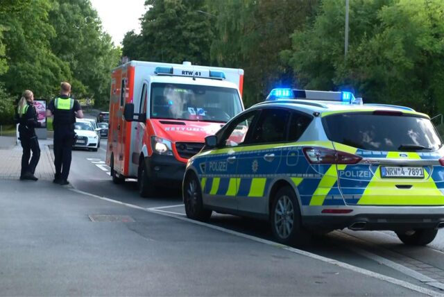 Ein Rettungswagen und ein Polizeiwagen stehen mit eingeschaltetem Blaulicht am Straßenrand. Zwei Polizisten stehen auf dem Gehweg.