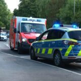 Ein Rettungswagen und ein Polizeiwagen stehen mit eingeschaltetem Blaulicht am Straßenrand. Zwei Polizisten stehen auf dem Gehweg.