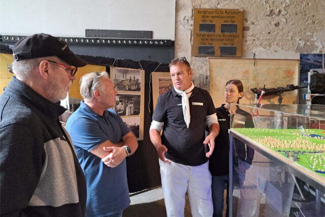 Eine Gruppe von Personen steht in einem Raum und betrachtet eine Ausstellung. Im Hintergrund sind eine Karte des Ruhrgebiets und Schilder mit Informationen zur Seilfahrt zu sehen.
