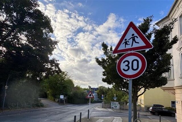 Ein Verkehrsschild mit der Aufschrift „30“ und einem Warnzeichen für Kinder befindet sich am Straßenrand. Die Geschwindigkeitsbegrenzung soll Verkehrsteilnehmer auf die mögliche Anwesenheit von spielenden Kindern hinweisen.