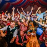 Männer und Frauen feiern in Trachten auf einem Oktoberfest. Sie stehen zum Teil auf den Bänken.