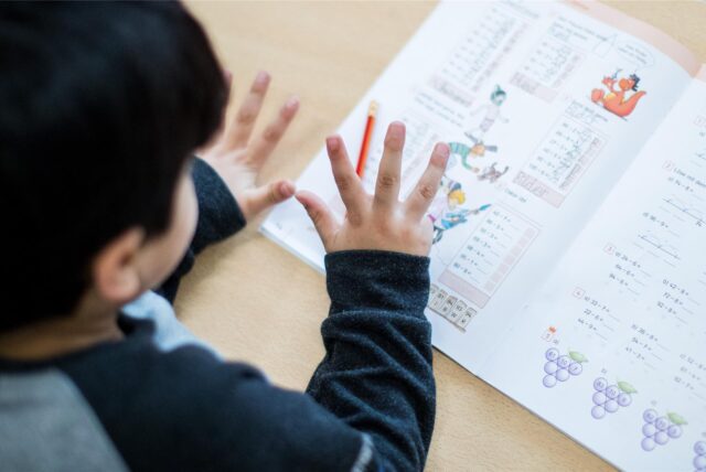 Ein Kind lernt mit einem Rechenbuch auf einem Tisch. Es scheint, als ob das Kind mit den Fingern zählt, während es die Aufgaben bearbeitet.