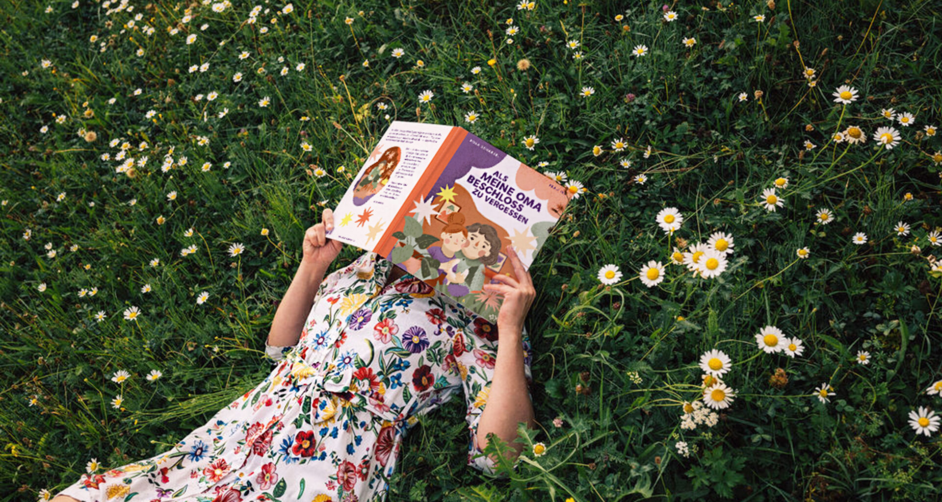 Frau mit sommerlichen Blümchenkleid liegt in der Wiese mit einem Buch vor dem Gesicht