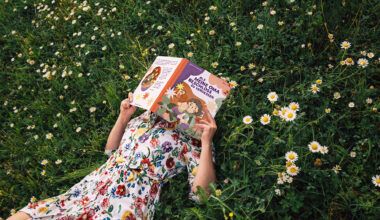 Frau mit sommerlichen Blümchenkleid liegt in der Wiese mit einem Buch vor dem Gesicht