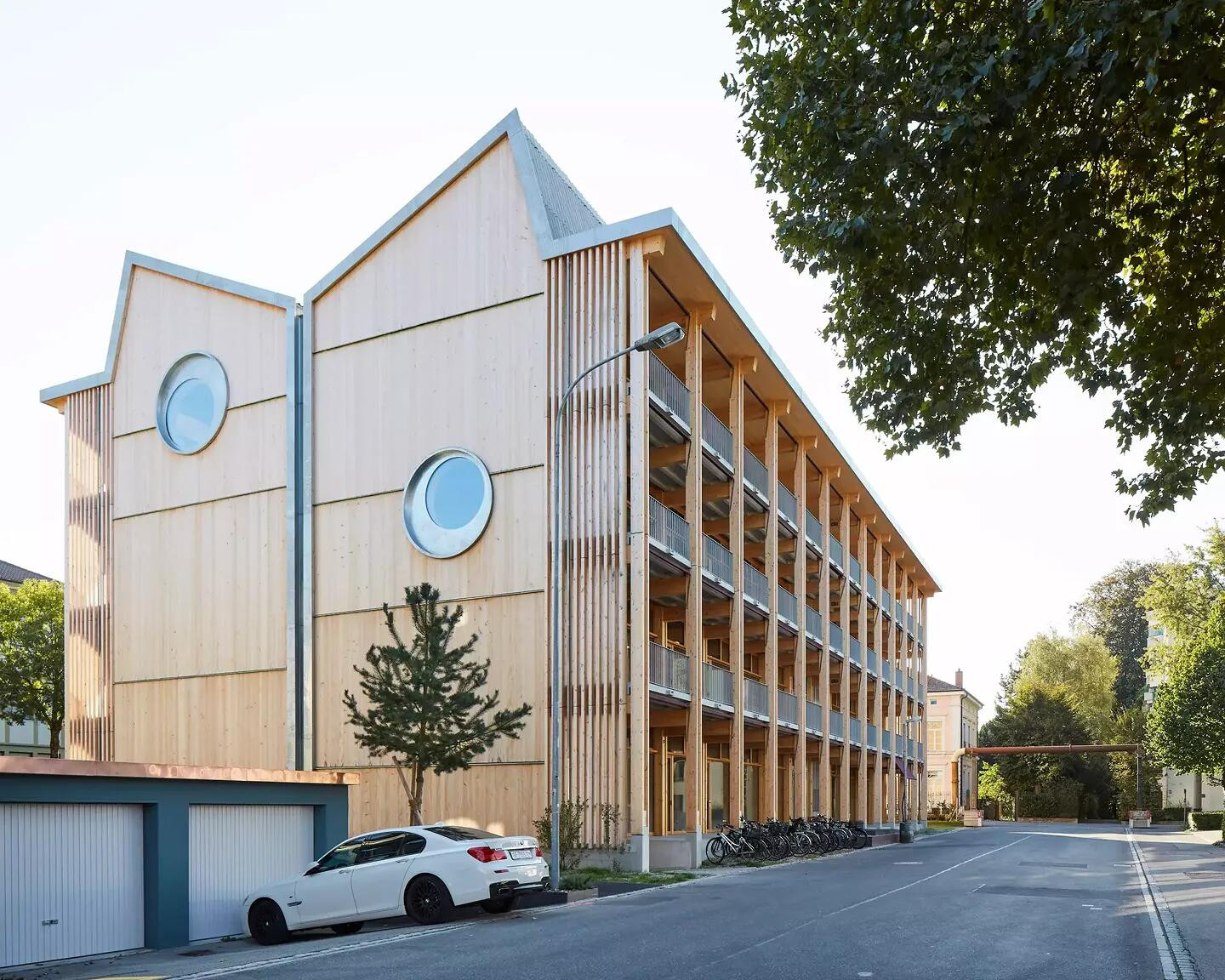 Dreistöckiges Holzgebäude mit runden Fenstern und überdachtem Balkon, umgeben von Bäumen und parkenden Autos.