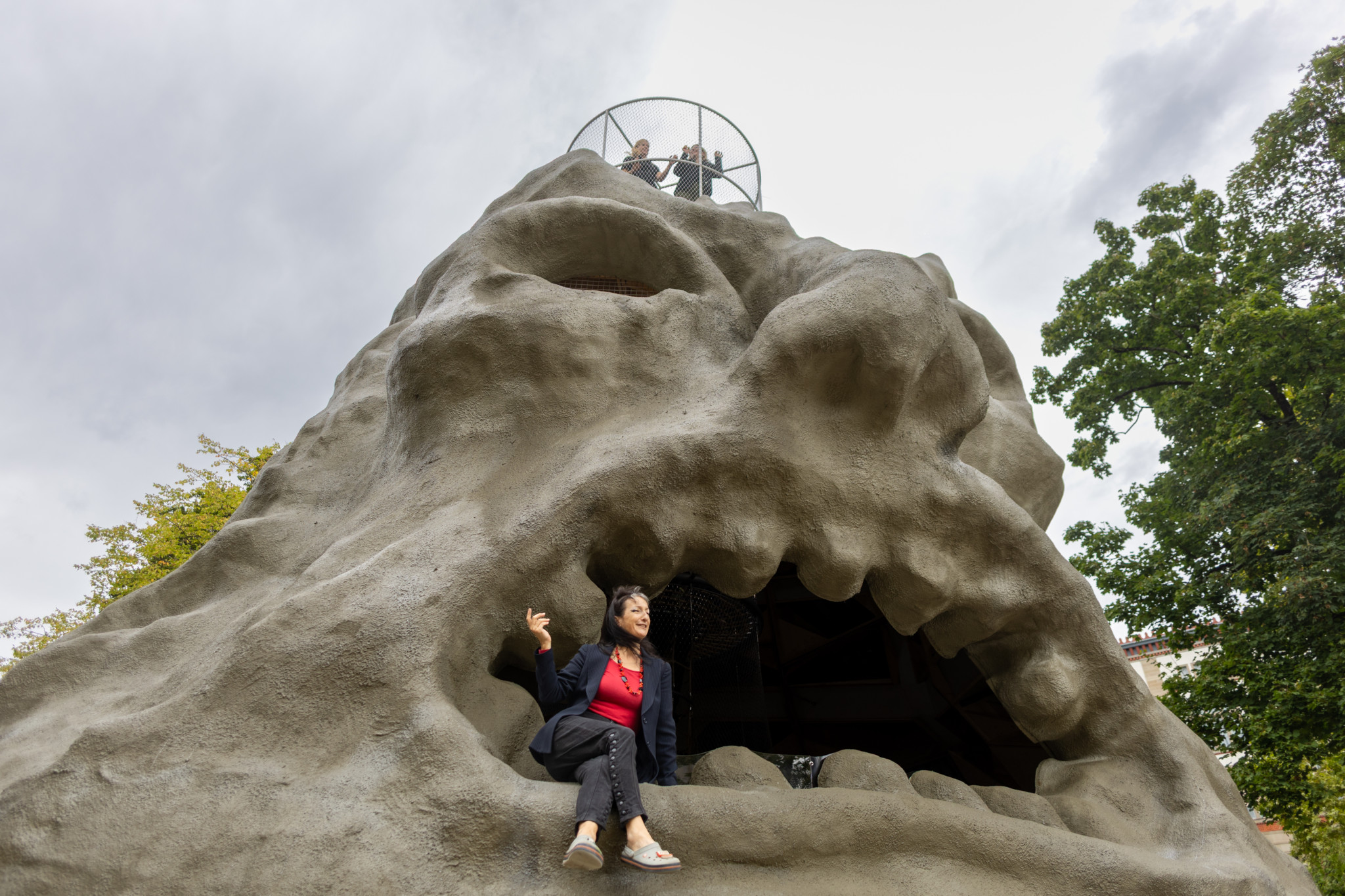 Das begehbare Kunstobjekt von Monster Chetwynd im Zürich Park zeigt eine Skulptur mit Klettergerüst, das an einen riesigen Kopf erinnert. Monster Chetwynd sitzt im geöffneten Mund der Skulptur, während zwei Menschen oben auf der Spitze stehen und den Ausblick geniessen.