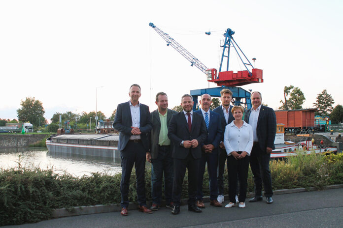 Eine Gruppe von acht Personen steht am Ufer eines Hafenbeckens und blickt in die Kamera. Hinter ihnen sind ein roter Portalkran, ein Frachtschiff am Kai sowie Gleisanlagen und Hafeninfrastruktur zu sehen. Abendlicht, Freiluftaufnahme