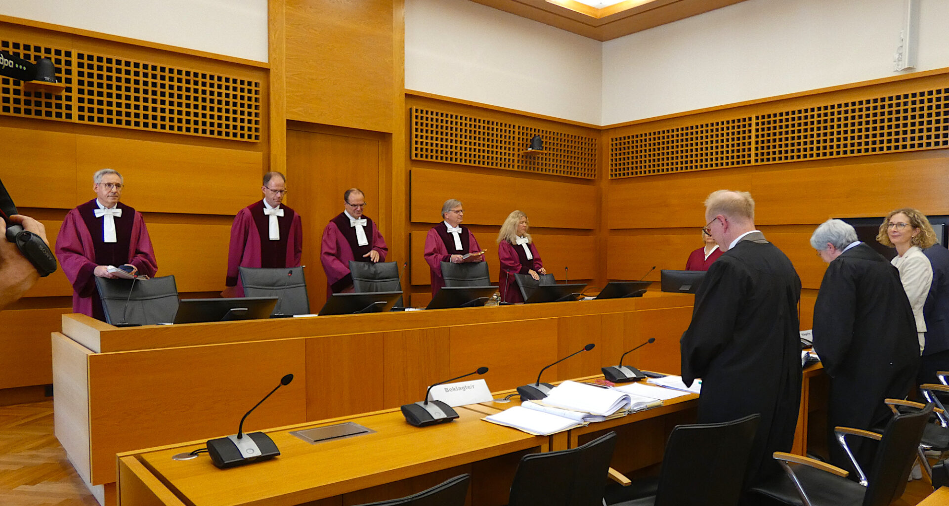 Richter in roten Roben stehen im Gerichtssaal hinter der Bank und schauen entgegen der Kamerarichtung.