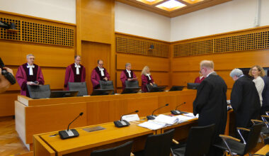 Richter in roten Roben stehen im Gerichtssaal hinter der Bank und schauen entgegen der Kamerarichtung.