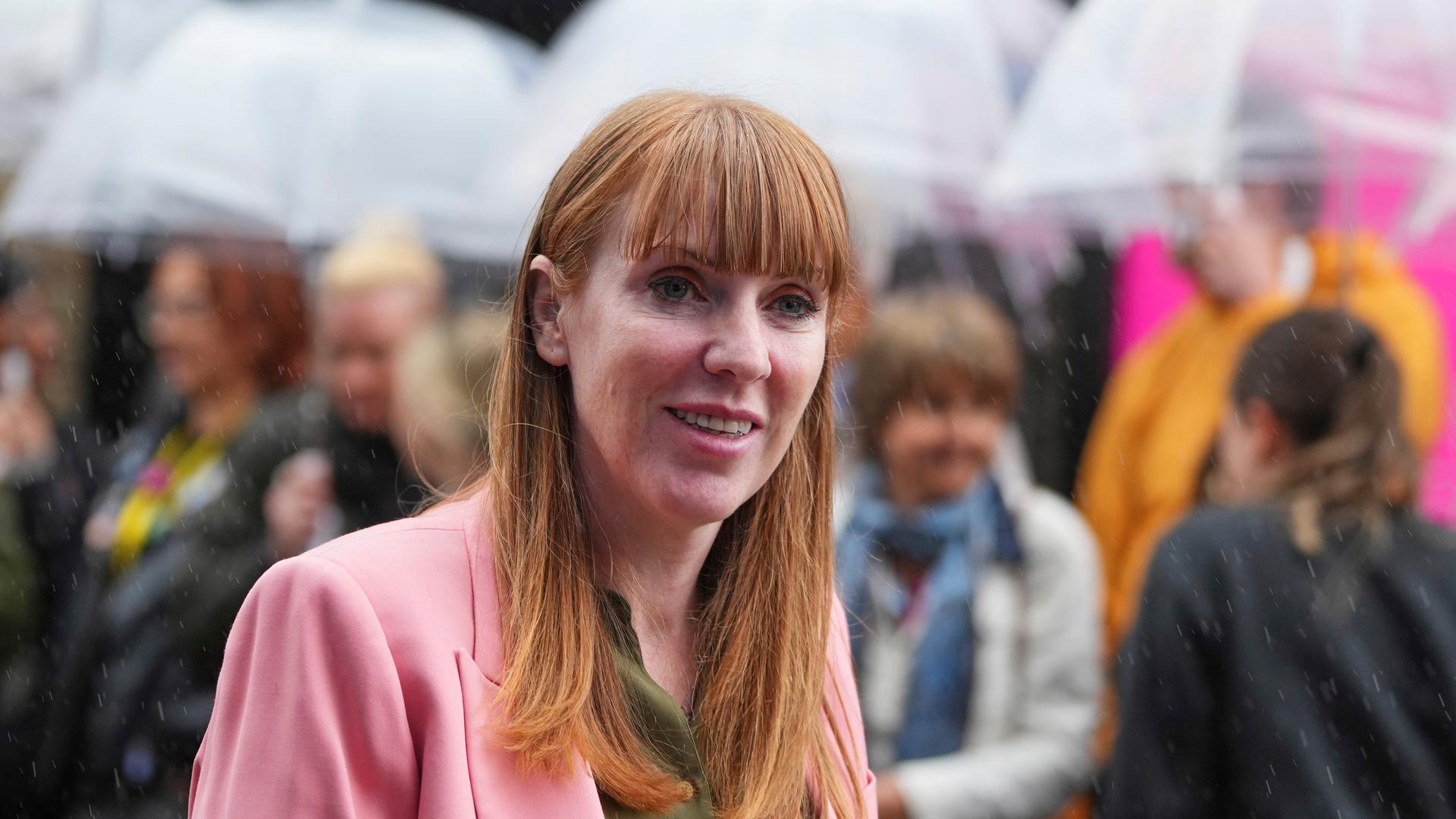 Angela Rayner lächelt in die Kamera. Hinter ihr sieht man verschwommen Menschen, die Regenschirme halten.
