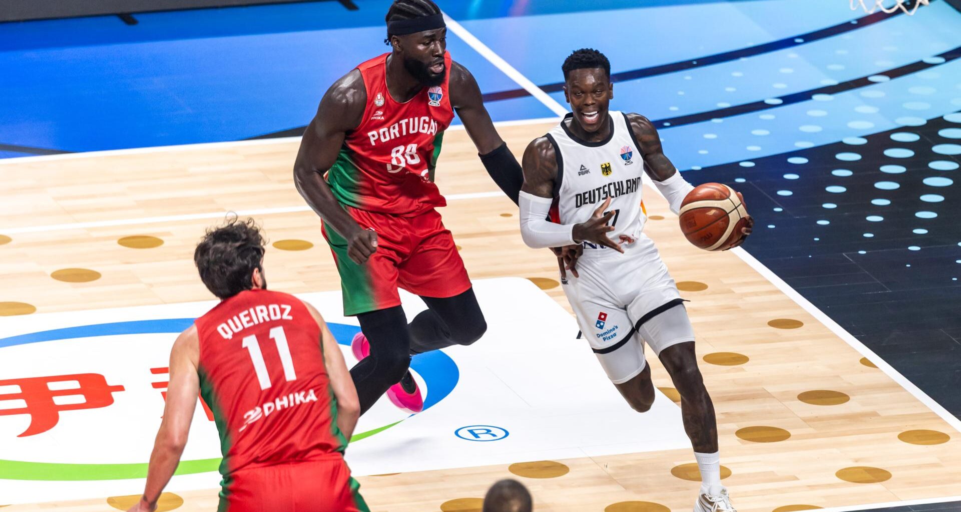 Basketballer Dennis Schröder dribbelt mit dem Ball und wird von einem Gegenspieler verfolgt.