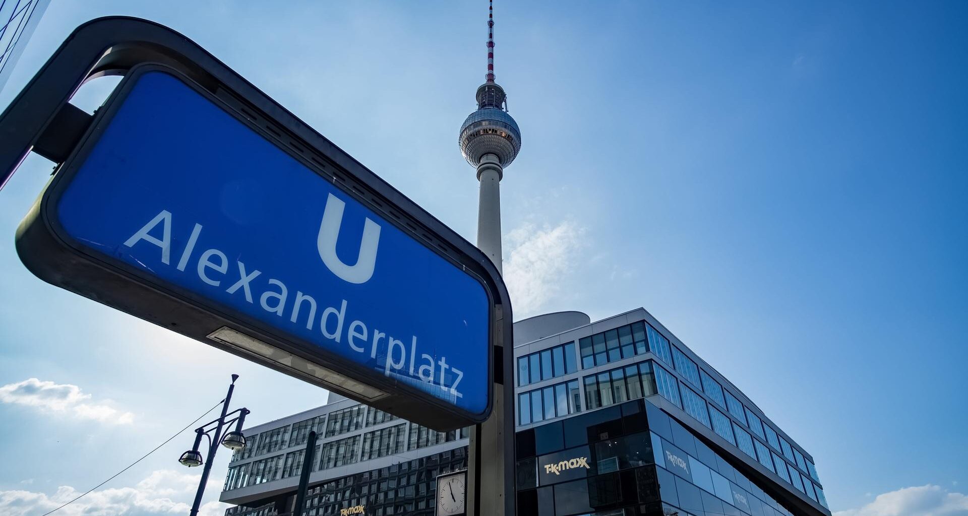 Aufnahme des U-Bahn Schildes am Bahnhof Alexanderplatz. Im Hintergrund ist vor blauem Himmel auch der Fernsehturm zu sehen.
