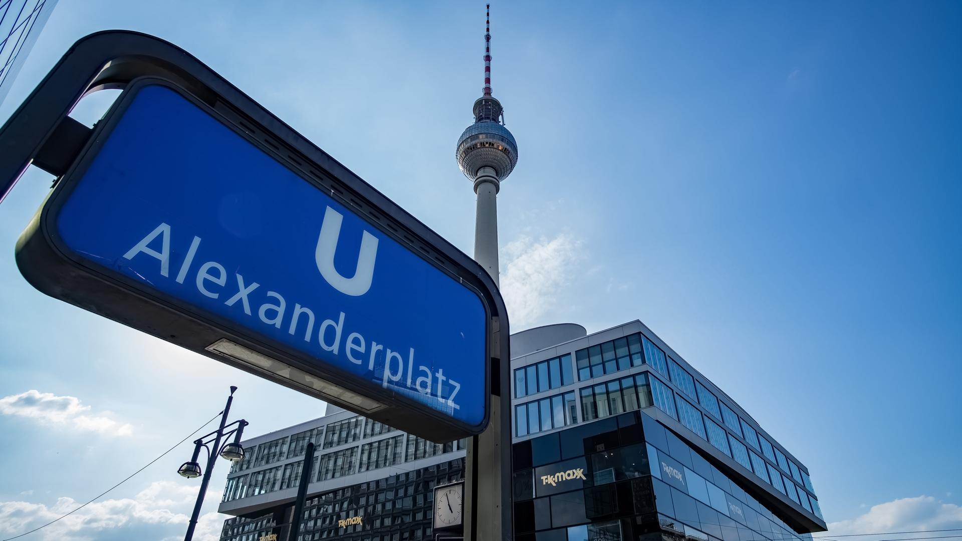 Aufnahme des U-Bahn Schildes am Bahnhof Alexanderplatz. Im Hintergrund ist vor blauem Himmel auch der Fernsehturm zu sehen.