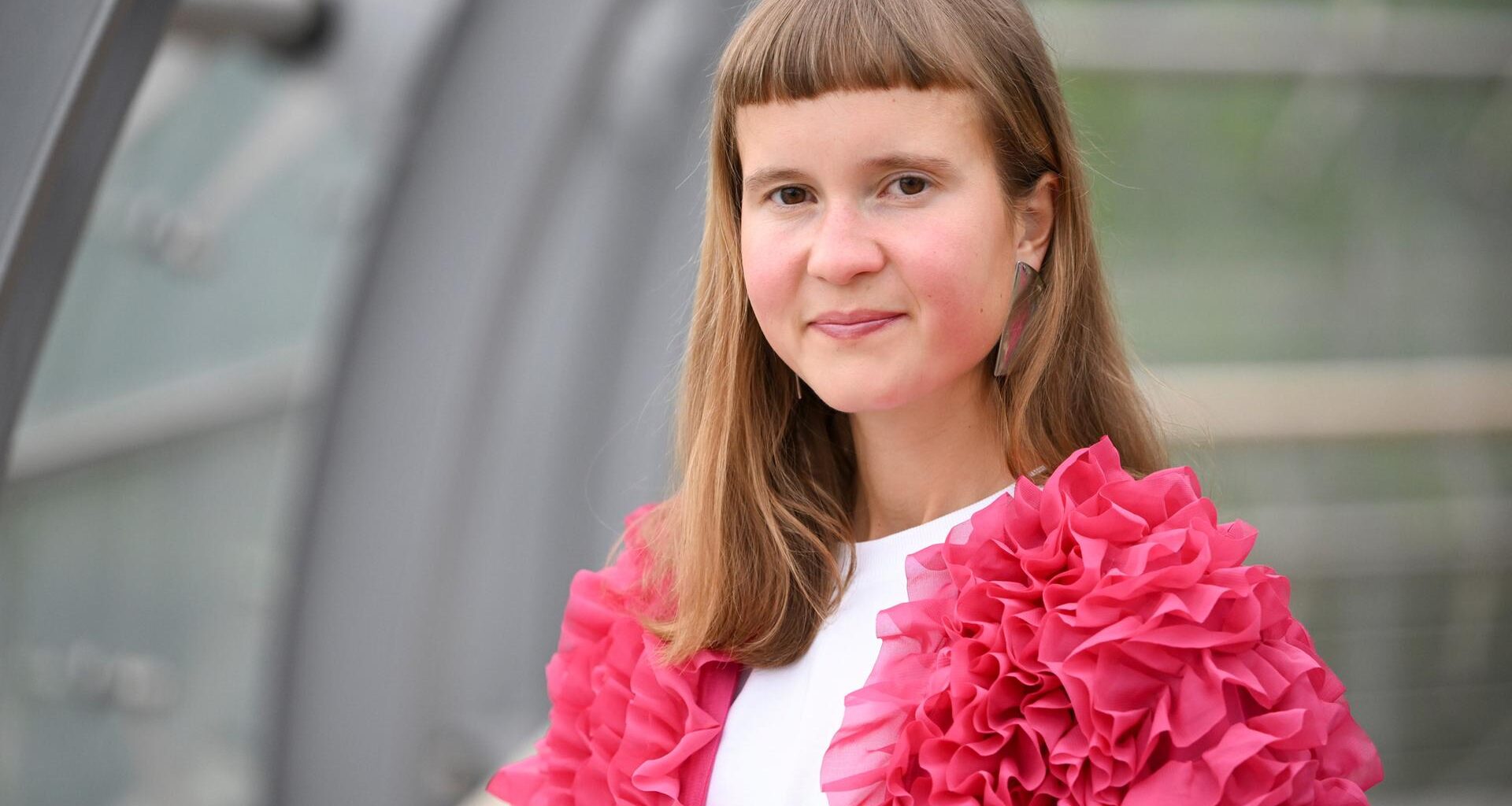Eine junge Frau mit ungewöhnlicher Pnyfrisur und einer pinken Jacke schaut in die Kamera: die Schriftstellerin Caroline Wahl