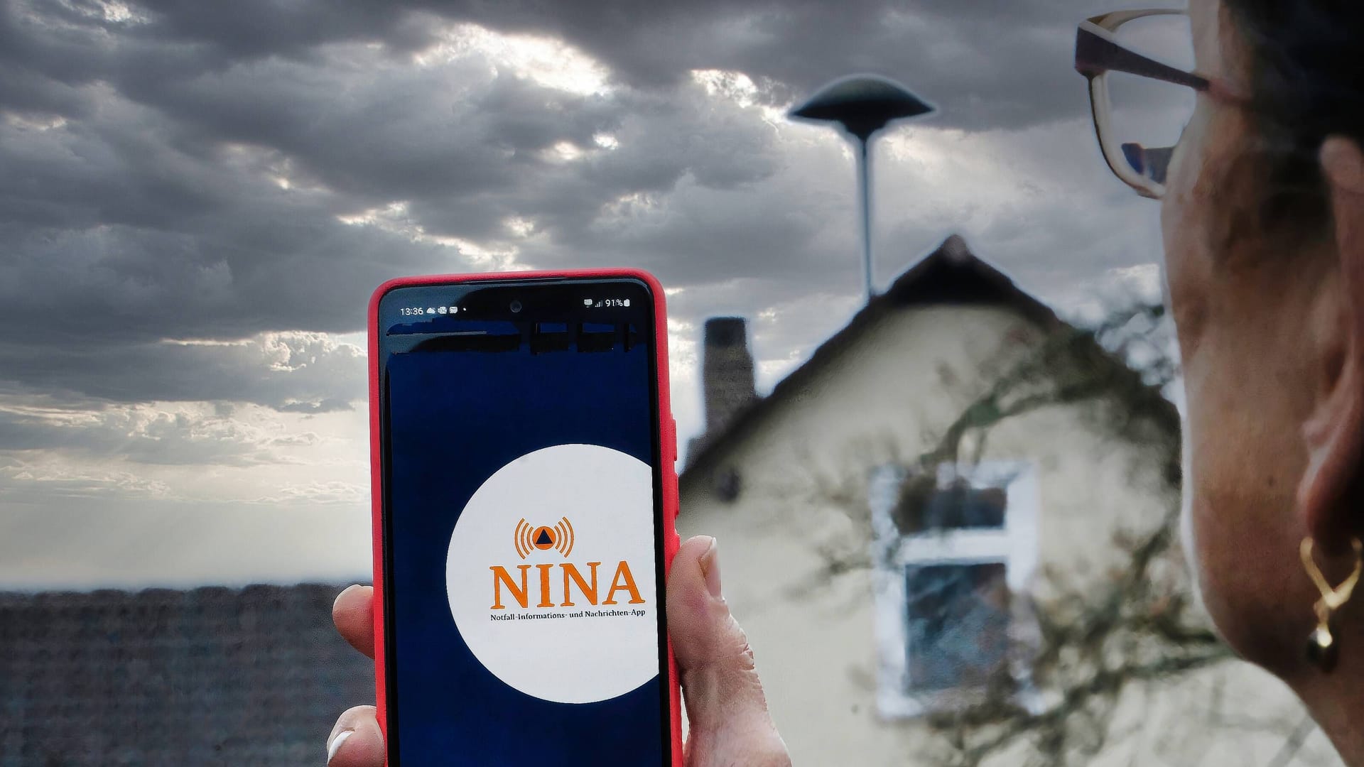 Eine Frau blickt auf eine Sirene und hat die Warn-App Nina auf ihrem Handy geöffnet (Symbolbild): Am Donnerstag geht der Warntag in die zweite Runde.