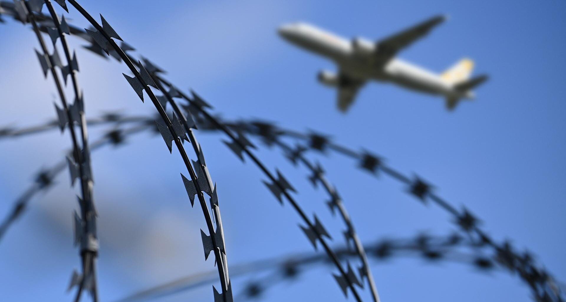 Ein startendes Passagierflugzeug ist am Himmel zu sehen, im Vordergrund ein Zaun mit Stacheldraht.